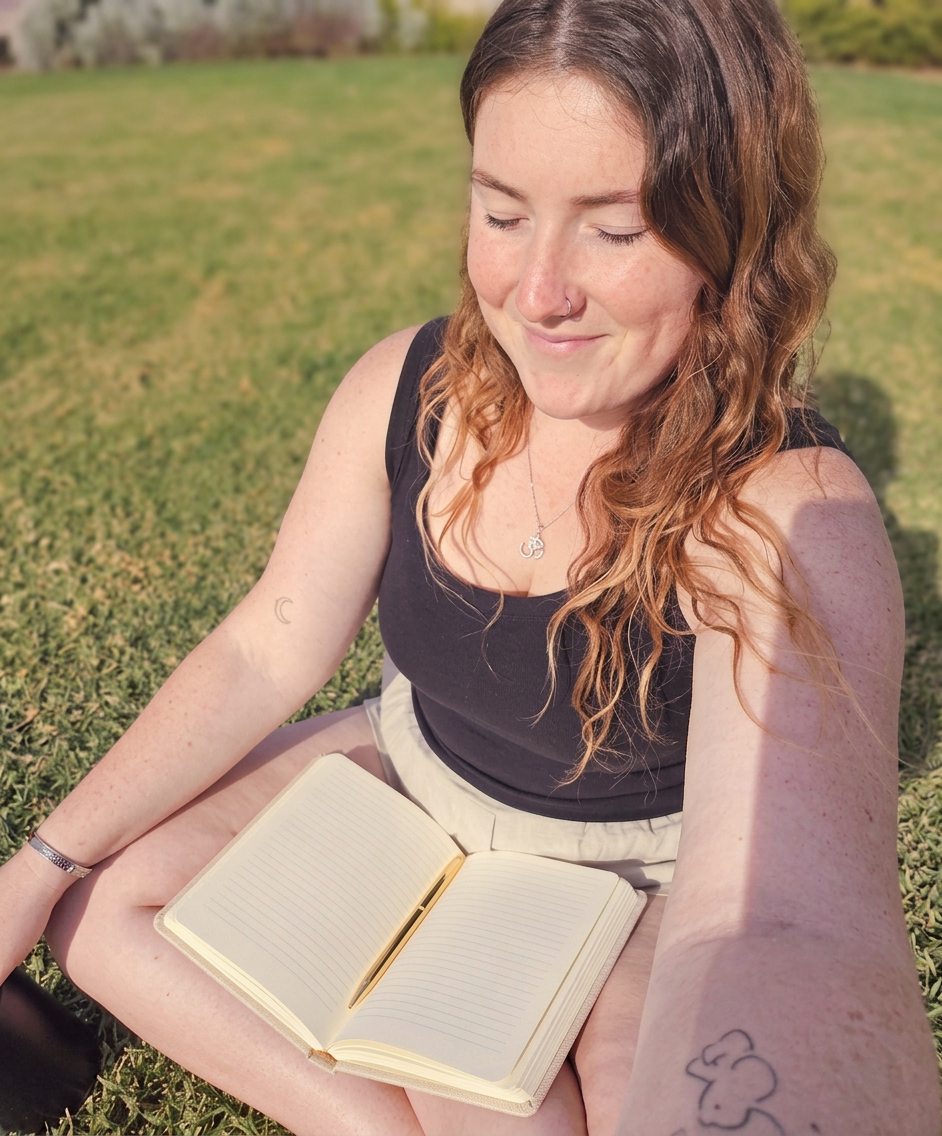 Caitie sitting on the grass with an open notebook in her lap, soft lavender light